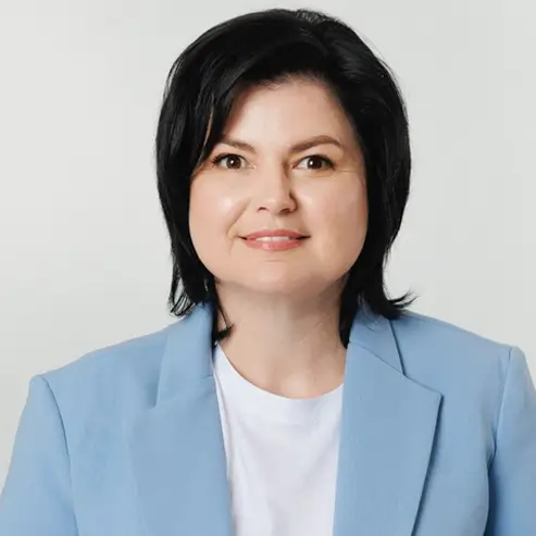 Засновниця магазину іграшок Тетяна Костовят, жінка з коротким темним волоссям, одягнена в світло-блакитний піджак поверх білого верху, на тлі світлого однотонного фону.