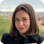 Засновниця магазину іграшок Анна Бисага, молода жінка з прямим темно-коричневим волоссям до плечей, одягнена в чорний жакет, стоїть на тлі пейзажу з деревами та будинками.